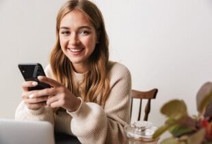 Young woman with long hair sits at a table, smiling and holding a smartphone, with a laptop and a glass jar in front of her, and a plant blurred in the foreground.