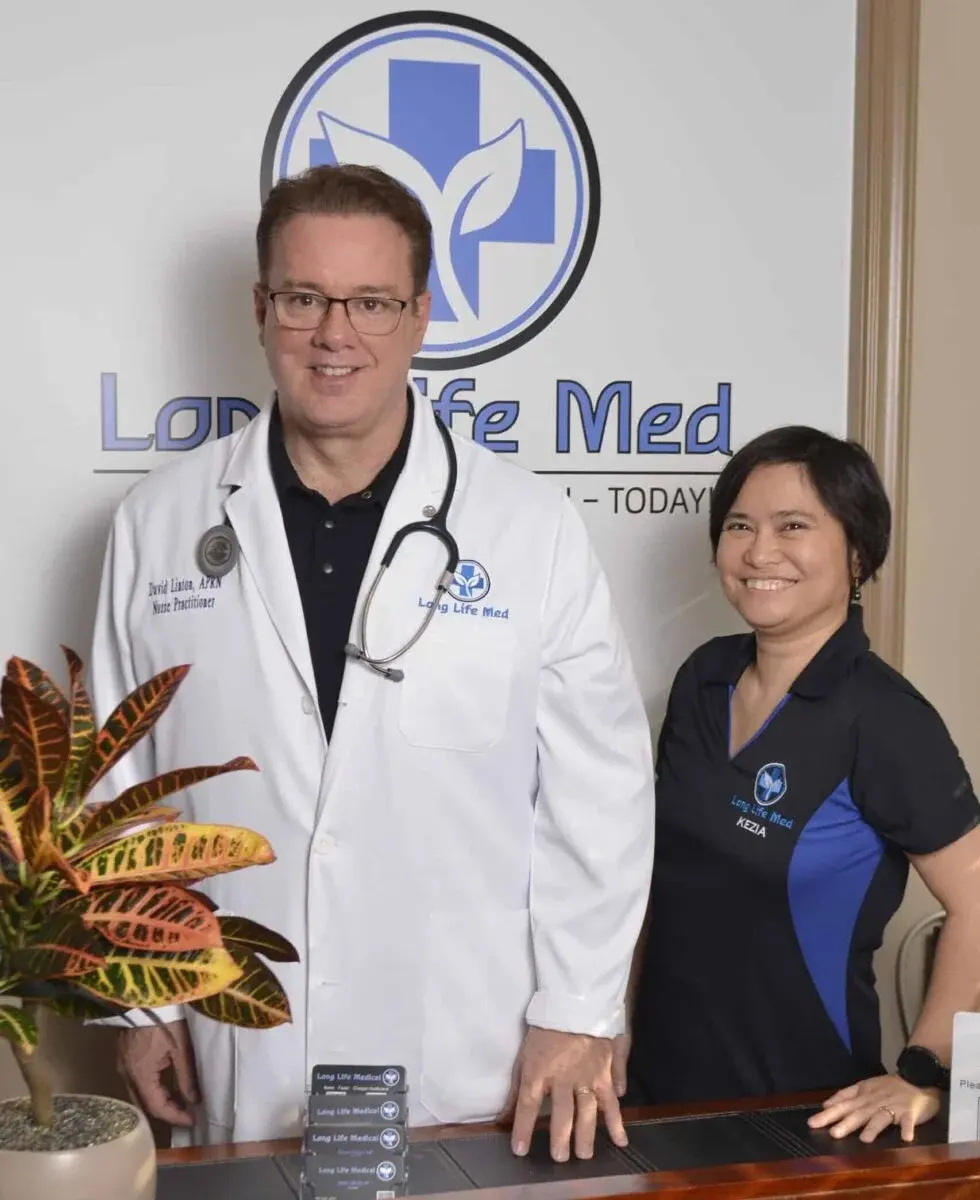 A man in a white medical coat and a woman in a black polo shirt stand at a reception desk in front of a "Long Life Med" sign.