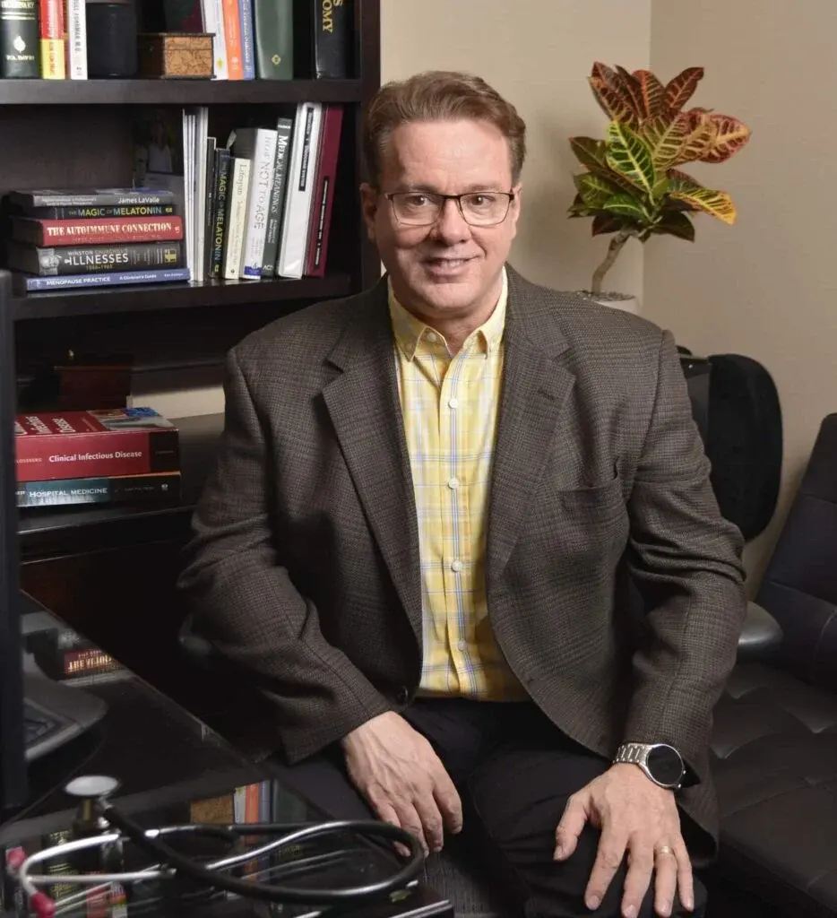 Man in glasses and a brown blazer sits in an office with bookshelves, a plant, and a stethoscope on the desk, reflecting his passion for helping others.