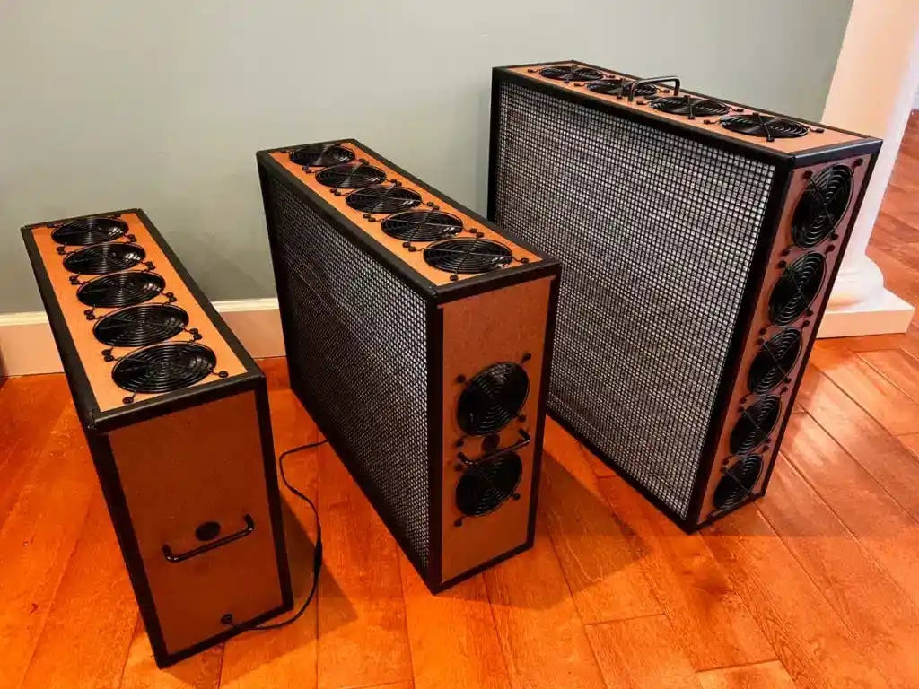 Three rectangular electronic devices with multiple cooling fans sit on a wooden floor next to a wall; one device is plugged in, reflecting our commitment to ensuring quality with every setup.